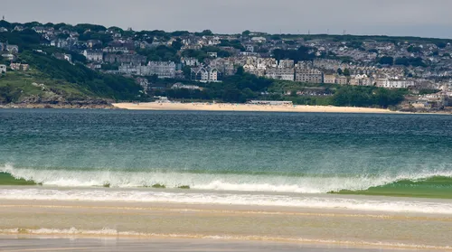 This is a landscape photograph showing Carbis Bay from Hayle in Cornwall, captured in the late morning during early summer. The image features the sea in the foreground, with gentle waves rolling onto the sandy beach. In the distance, the town of St Ives is visible, with its distinctive array of houses and buildings spread across the hillside. The scene captures the coastal beauty of Cornwall, highlighting the relationship between the sea, the expansive beach, and the urban area of St Ives.